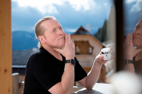 Joey Kelly sitzt an einem Holztisch, in der Hand ein Körperpflegeprodukt. Er trägt ein schwarzes T-Shirt und eine Armbanduhr. Im Hintergrund blauer Himmel.