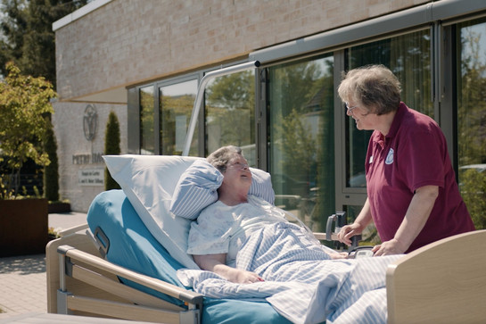 Eine Frau liegt vor dem Hospiz in einem Bett und unterhält sich.