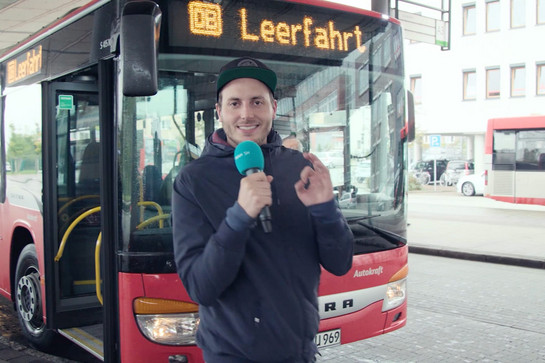 bb_helmut_teaser_04_hauptteaser.jpg Ein Mann steht mit seinem Mikro vor einem Bus