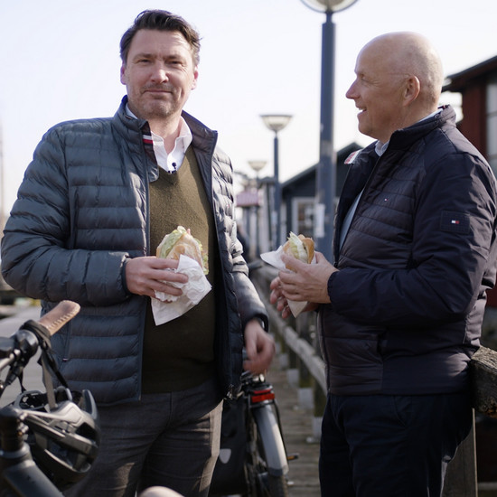 Clip für Wohnungsbau-gesellschaft Quadratisches Bild von zwei Herren, die jeweils ein Fischbrötchen am Hafen essen