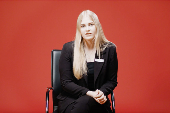 Eine junge Frau sitzt vor einer roten Wand auf einem Bürostuhl 