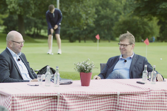bb_stegner_teaser_03.jpg Zwei Männer sitzen an einem Tisch der auf einem Golfplatz sitzt