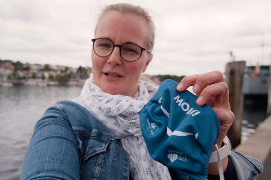 Eine blonde Frau hält eine Maske in die Kamera