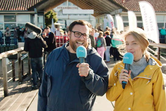 bb_hotdog_teaser_01.jpg Zwei Journalisten stehen mit Mikrofonen an einem Steg