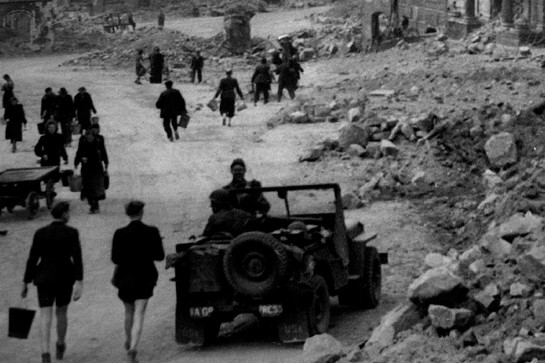 Historische Aufnahme von Personen und Auto neben Trümmern