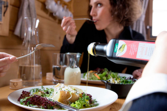 Zwei Personen sitzen an einem Tisch, sie essen Salat mit Ofenkartoffeln. Jemand tut etwas Speiseöl auf den Salat, auf der Flasche steht „Cellagon“.