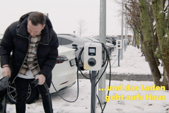 Mann an einer Ladestation. Gelber Slogan: „… und das Laden geht aufs Haus“