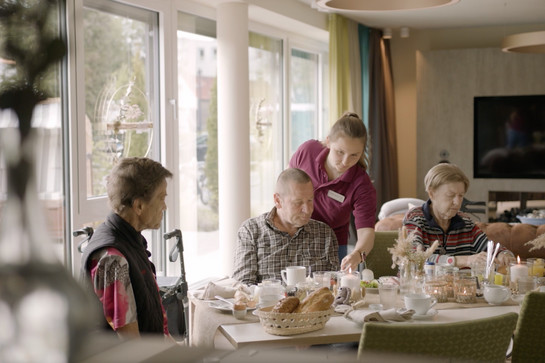 Drei ältere Menschen sitzen am Frühstückstisch und werden von einer Pflegerin betreut