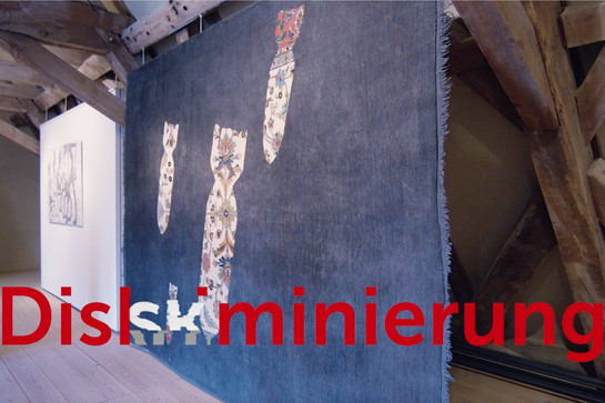 Kunstwerk aus Stoff mit fallenden Bomben. Darunter roter Text: "Diskriminierung"