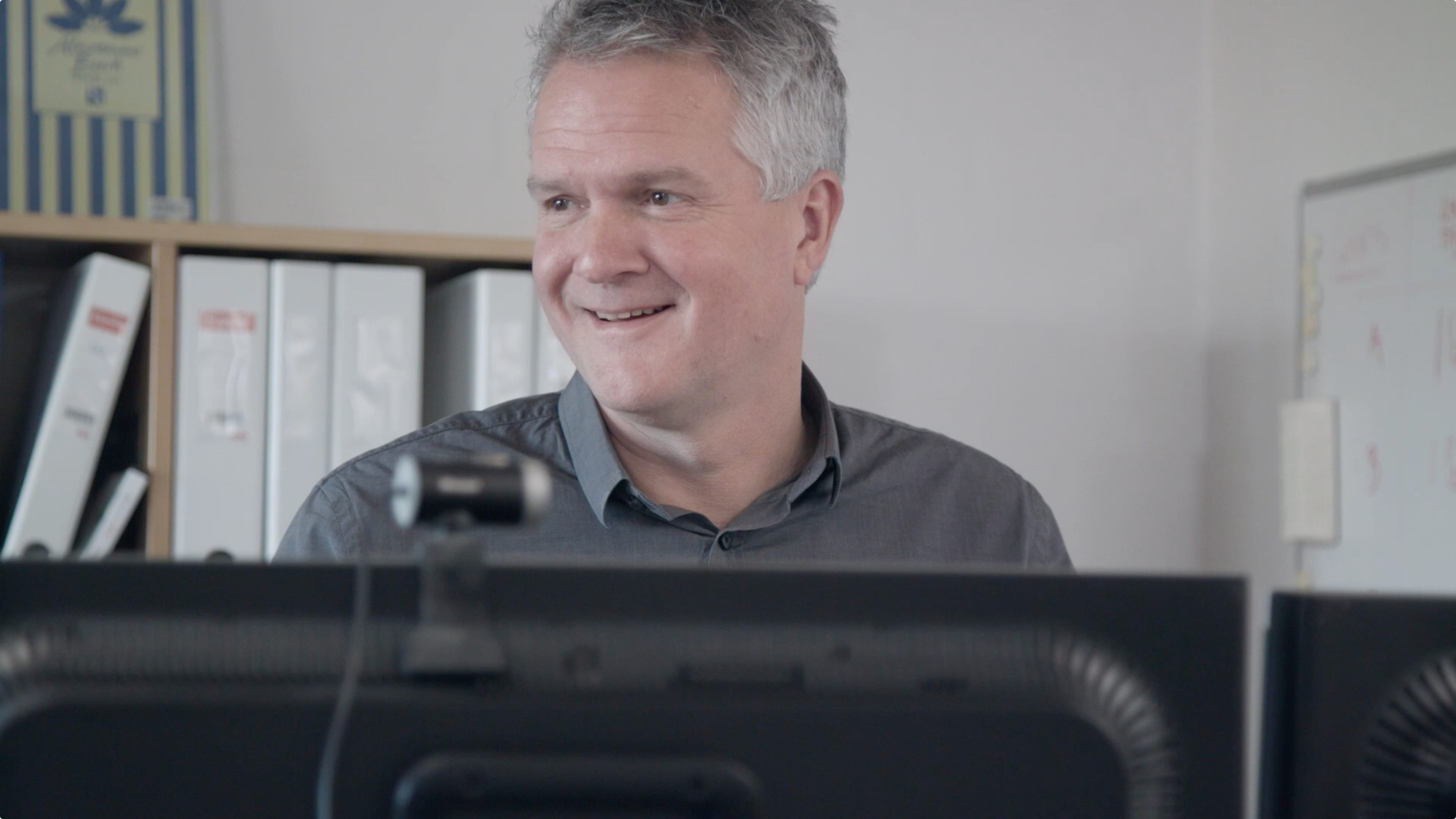 An einem Schreibtisch sitzt ein Mann mit grauen Haaren an einem Computer. 