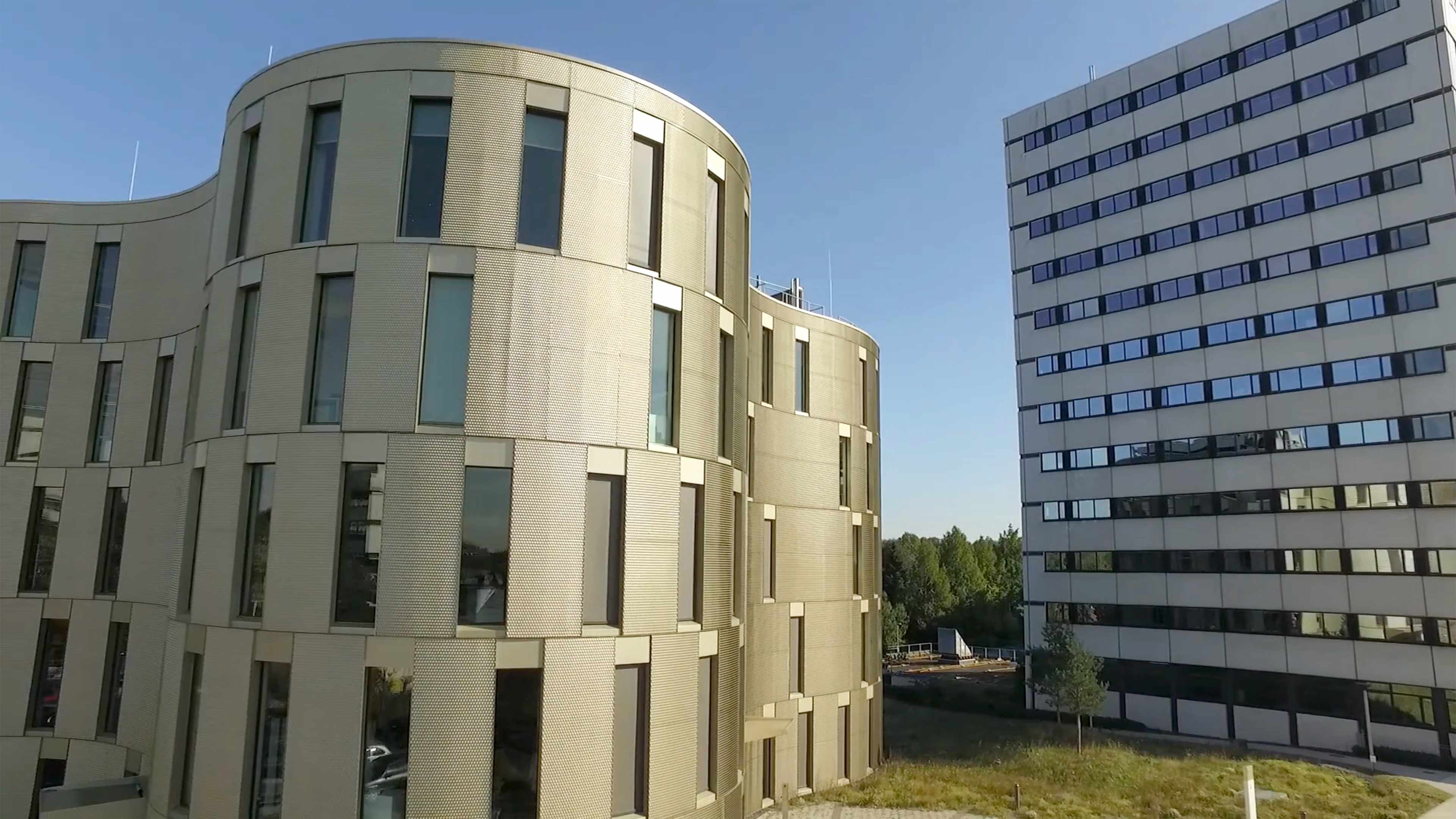 Zwei große Gebäude, das eine hat rundliche Formen. Das andere ist eckig. Beide sind grau und haben viele Fenster. 