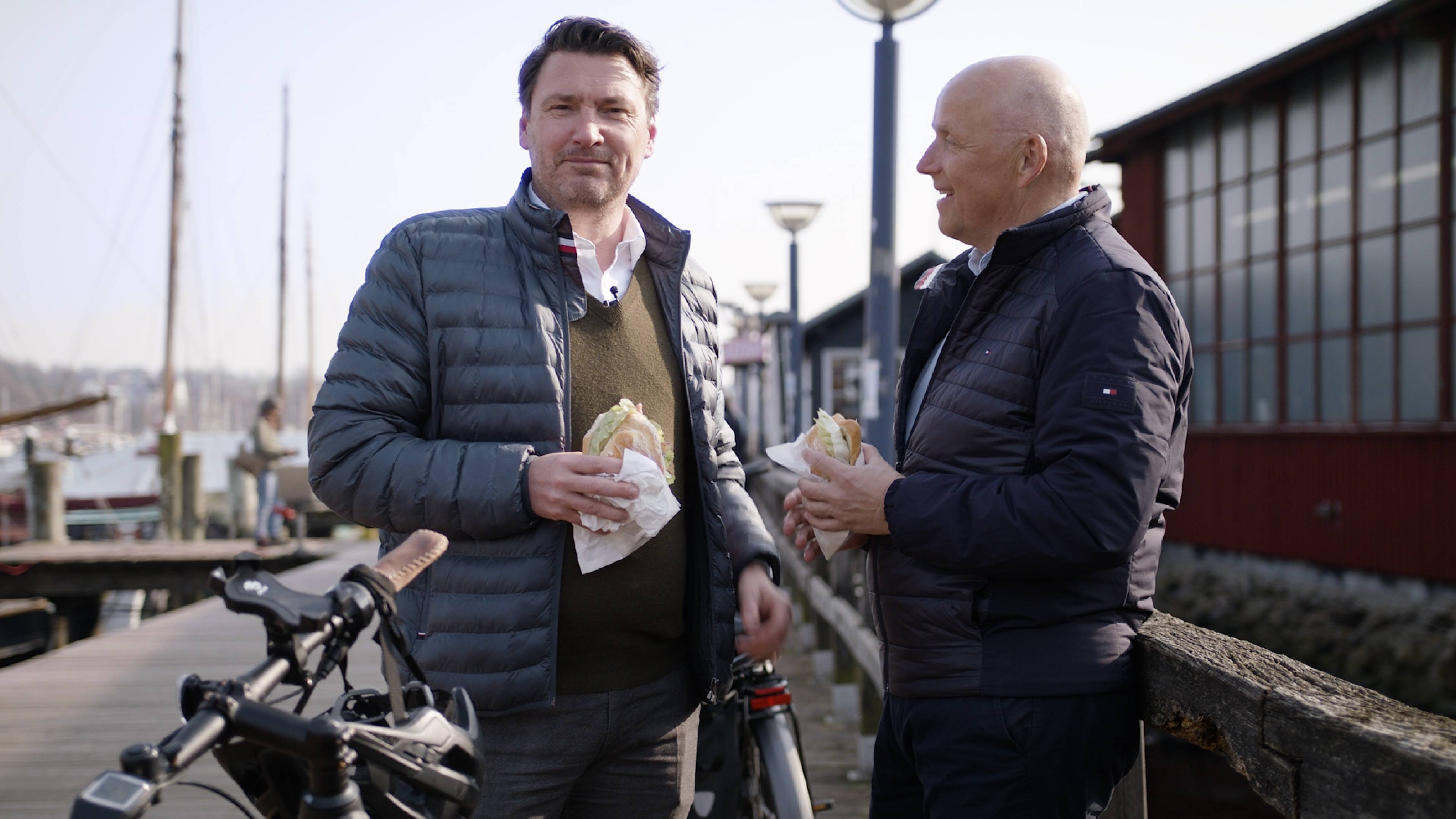 Rechteckiges Bild von zwei Herren, die jeweils ein Fischbrötchen am Hafen essen